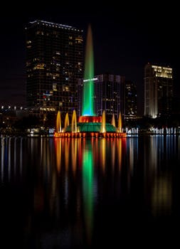 Illuminated fountain at night with Orlando skyline backdrop, reflecting vibrant lights on Lake Eola.