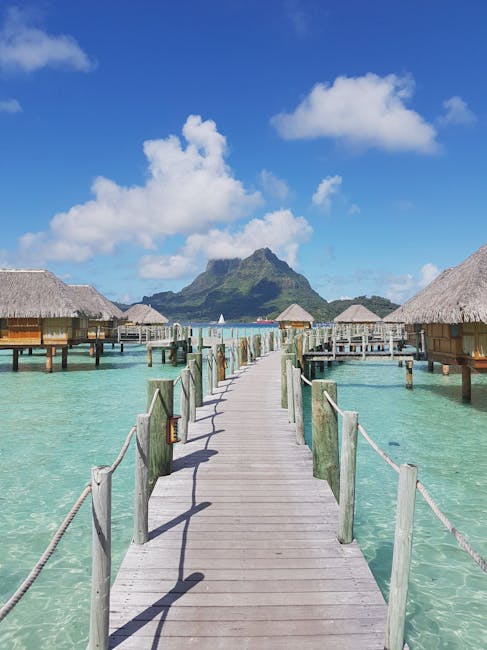 Idyllic tropical scene with wooden walkways and overwater bungalows overlooking a mountain on a sunny day.