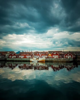 Picturesque view of a waterfront in Vågan, Norway with red cabins and a boat reflecting on calm waters under a dramatic sky.
