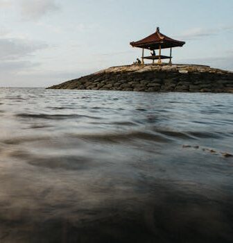 Tranquil scene of a traditional pagoda on a rocky island in Bali's serene sunset waters.